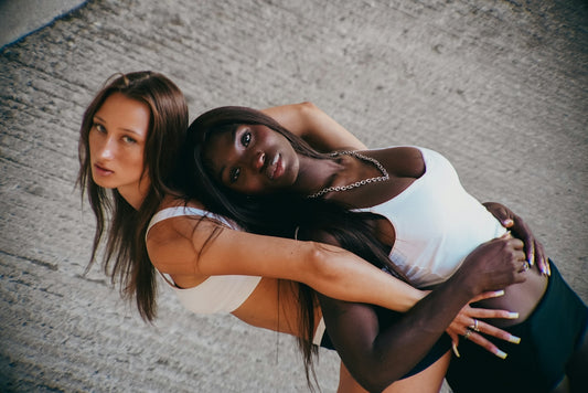 Two women in white tank tops leaning back to back against a concrete wall